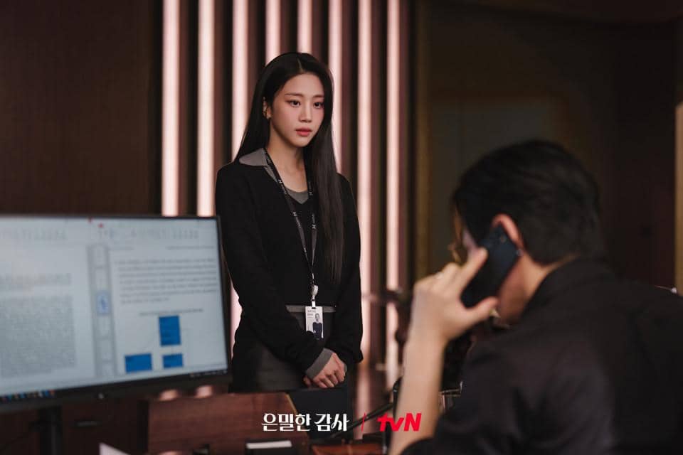 Seorang wanita berpakaian formal berdiri di depan meja kerja dengan komputer, sementara seorang pria berbicara di telepon di ruang kantor modern.