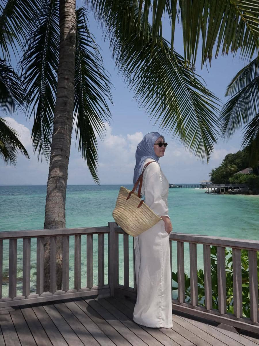 Seorang wanita berhijab mengenakan gaun putih dan kacamata hitam berdiri di tepi dermaga kayu dengan latar laut biru dan pohon kelapa.
