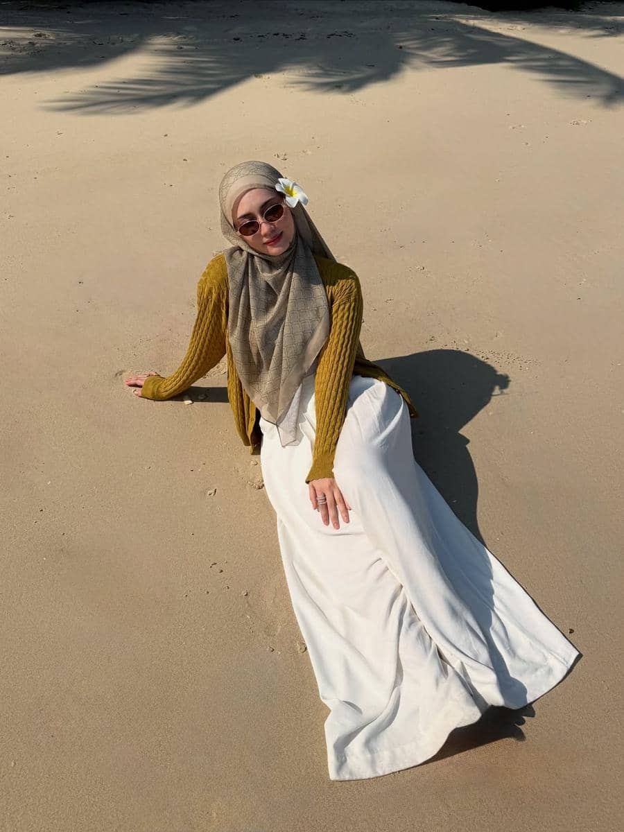 Seorang wanita berhijab mengenakan kacamata hitam, sweater mustard, dan rok putih duduk santai di pasir pantai dengan bunga di hijabnya.
