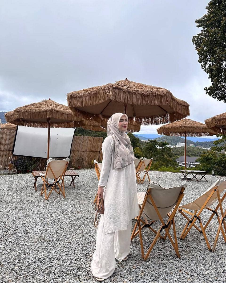 Seorang wanita berhijab mengenakan busana serba putih bergaya santai berdiri di area outdoor dengan kursi lipat dan payung jerami.