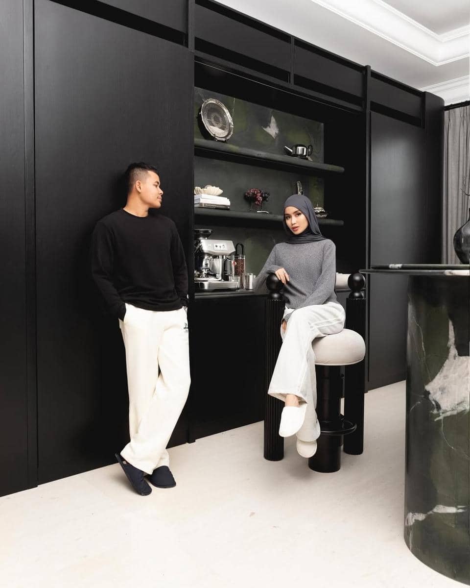 Sepasang pria dan wanita berbusana kasual monokrom berpose di dapur modern dengan latar perabot hitam elegan dan mesin kopi.