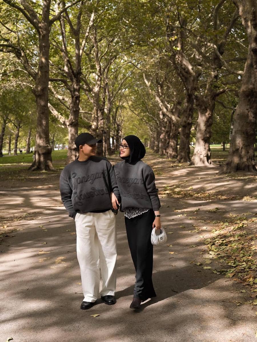 Sepasang pasangan berjalan bersama di jalan taman rindang dengan mengenakan sweater abu-abu senada dan tampak saling tersenyum.