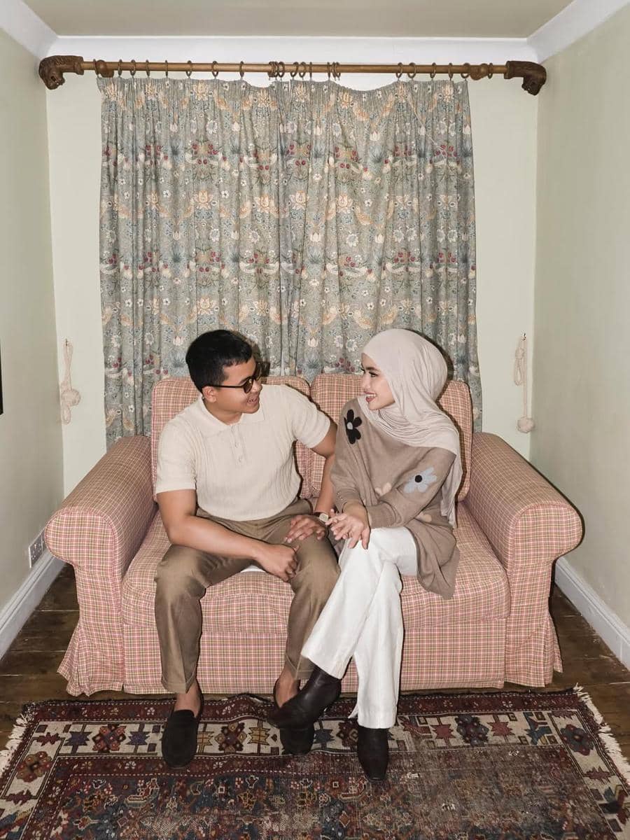 Sepasang pria dan wanita duduk di sofa bermotif kotak, mengenakan busana kasual bernuansa cokelat dan krem dengan latar tirai bermotif bunga.