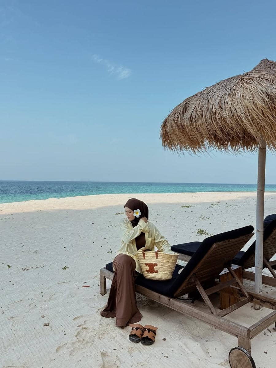 Seorang wanita berhijab duduk di kursi pantai kayu di bawah payung jerami, mengenakan pakaian santai dengan tas anyaman di tepi pantai berpasir putih.
