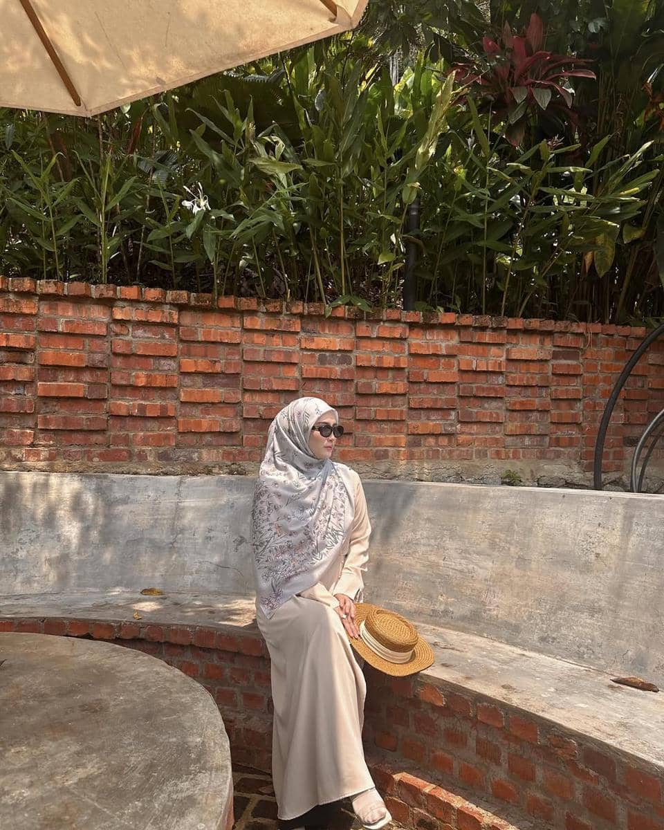 Seorang wanita berhijab duduk di bangku semen dengan latar dinding bata merah, mengenakan gaun krem dan memegang topi jerami.