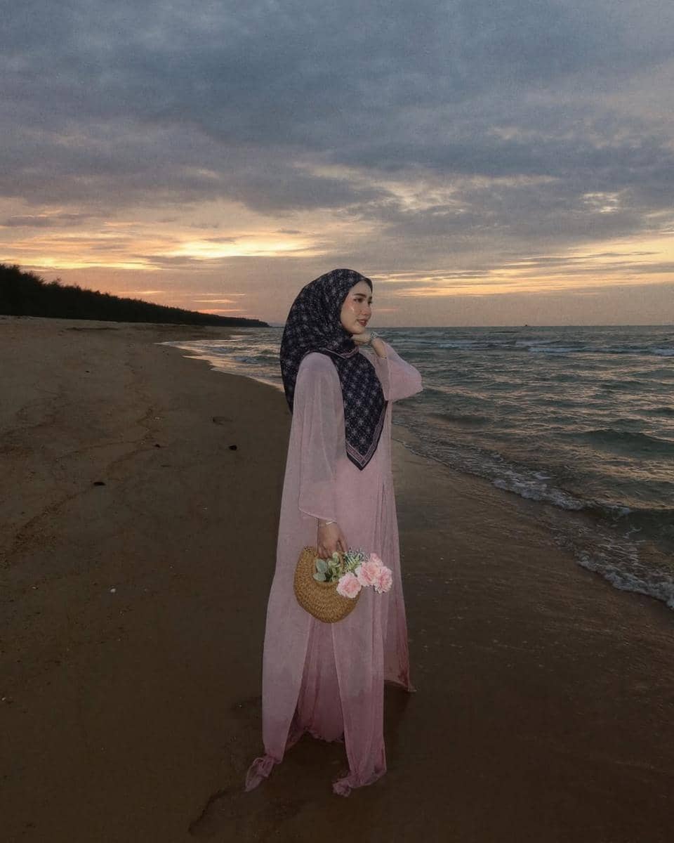 Seorang wanita berhijab mengenakan gaun panjang berwarna pink sambil memegang tas rotan berisi bunga di tepi pantai saat senja.