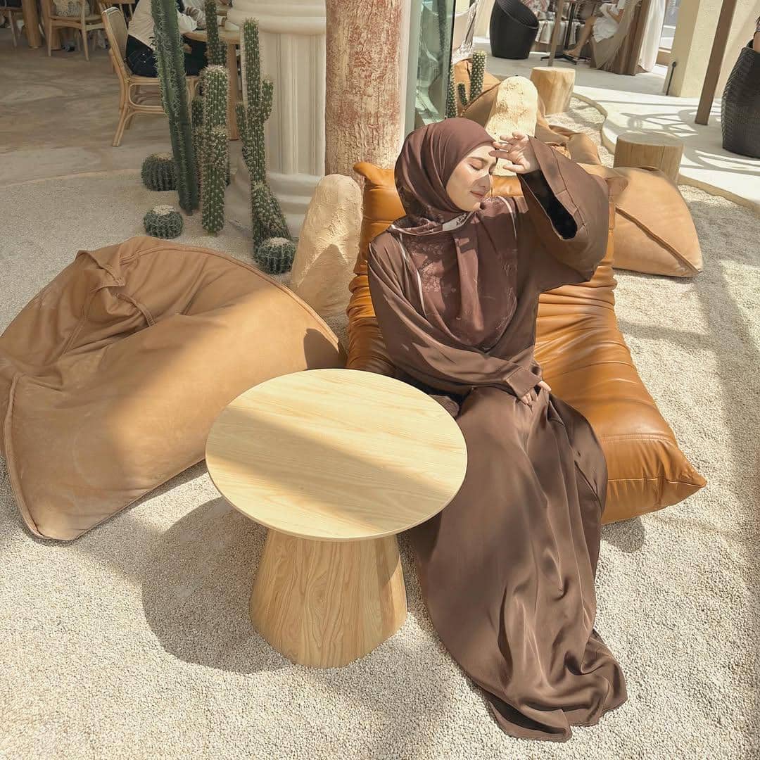 Seorang wanita berhijab cokelat duduk santai di kursi bean bag berwarna cokelat di area bernuansa pantai dengan dekorasi kaktus.