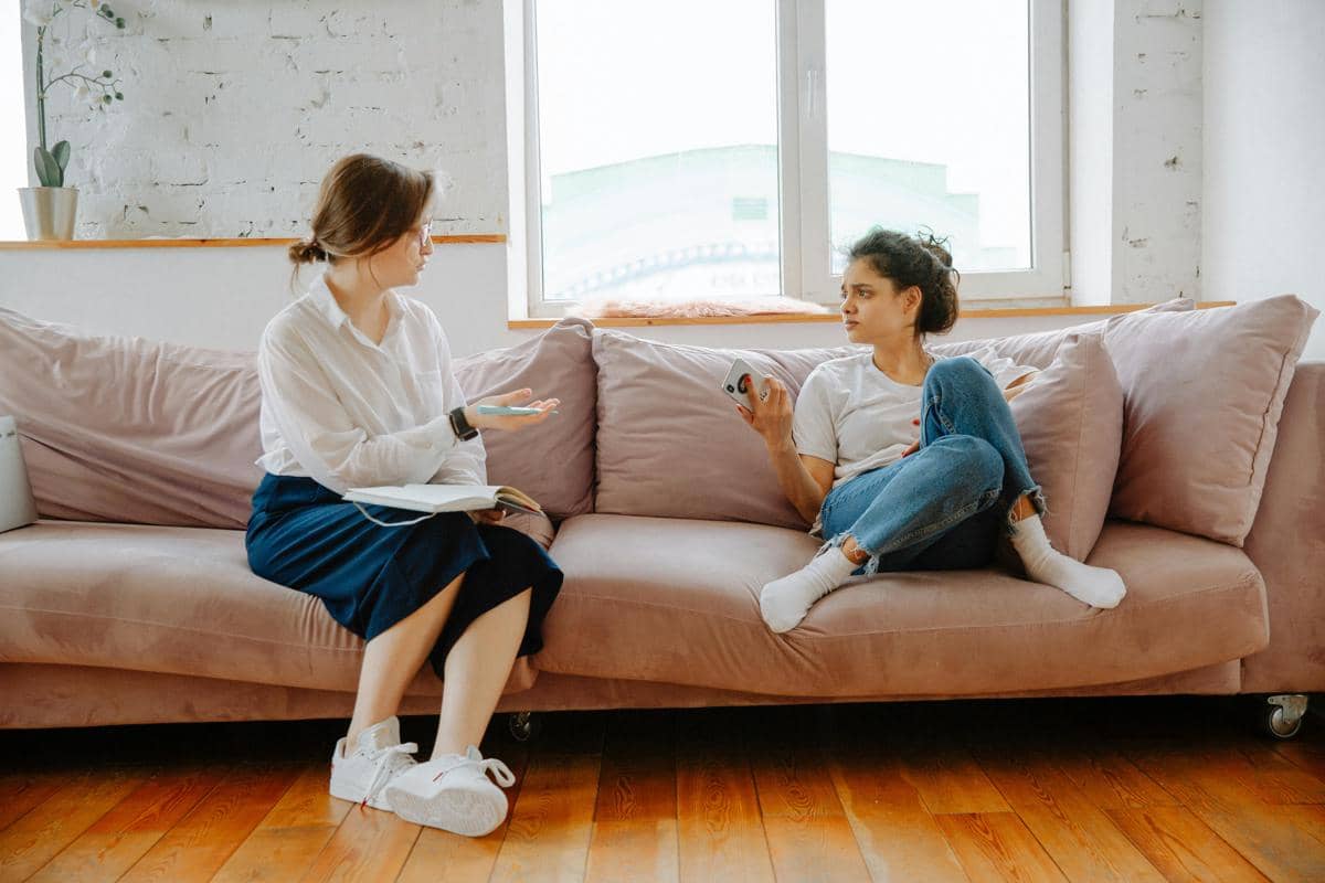 Dua orang duduk di sofa berwarna lembut sedang berbincang serius di ruang tamu dengan cahaya alami dari jendela besar.