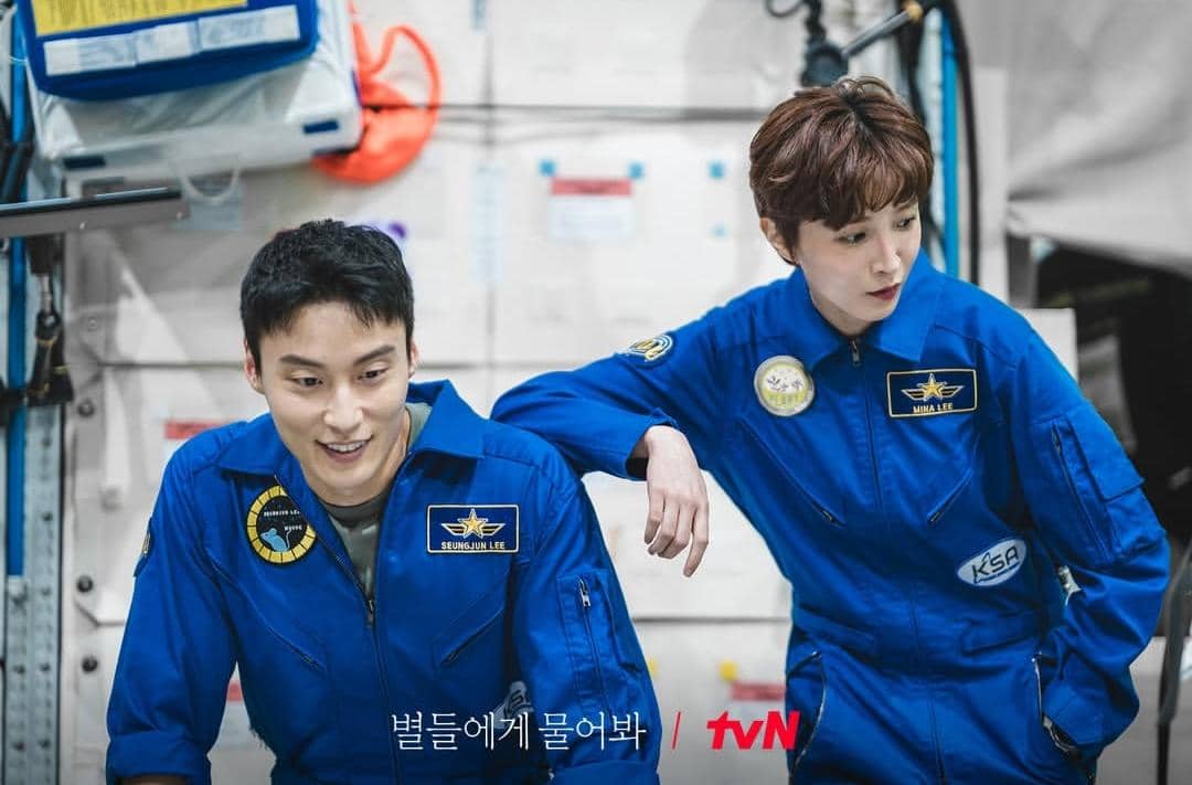 Dua aktor mengenakan seragam astronot biru dengan nama di dada, berada di dalam ruangan yang tampak seperti fasilitas pelatihan luar angkasa.