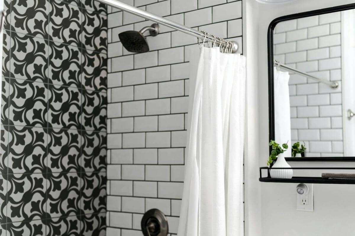Kamar mandi bergaya modern dengan dinding ubin putih dan hitam, tirai shower putih, serta cermin dengan bingkai hitam.
