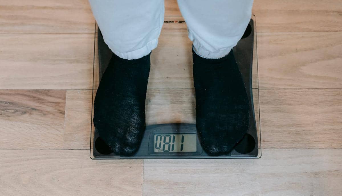 Seseorang mengenakan kaus kaki hitam berdiri di atas timbangan digital di lantai kayu untuk mengukur berat badan.