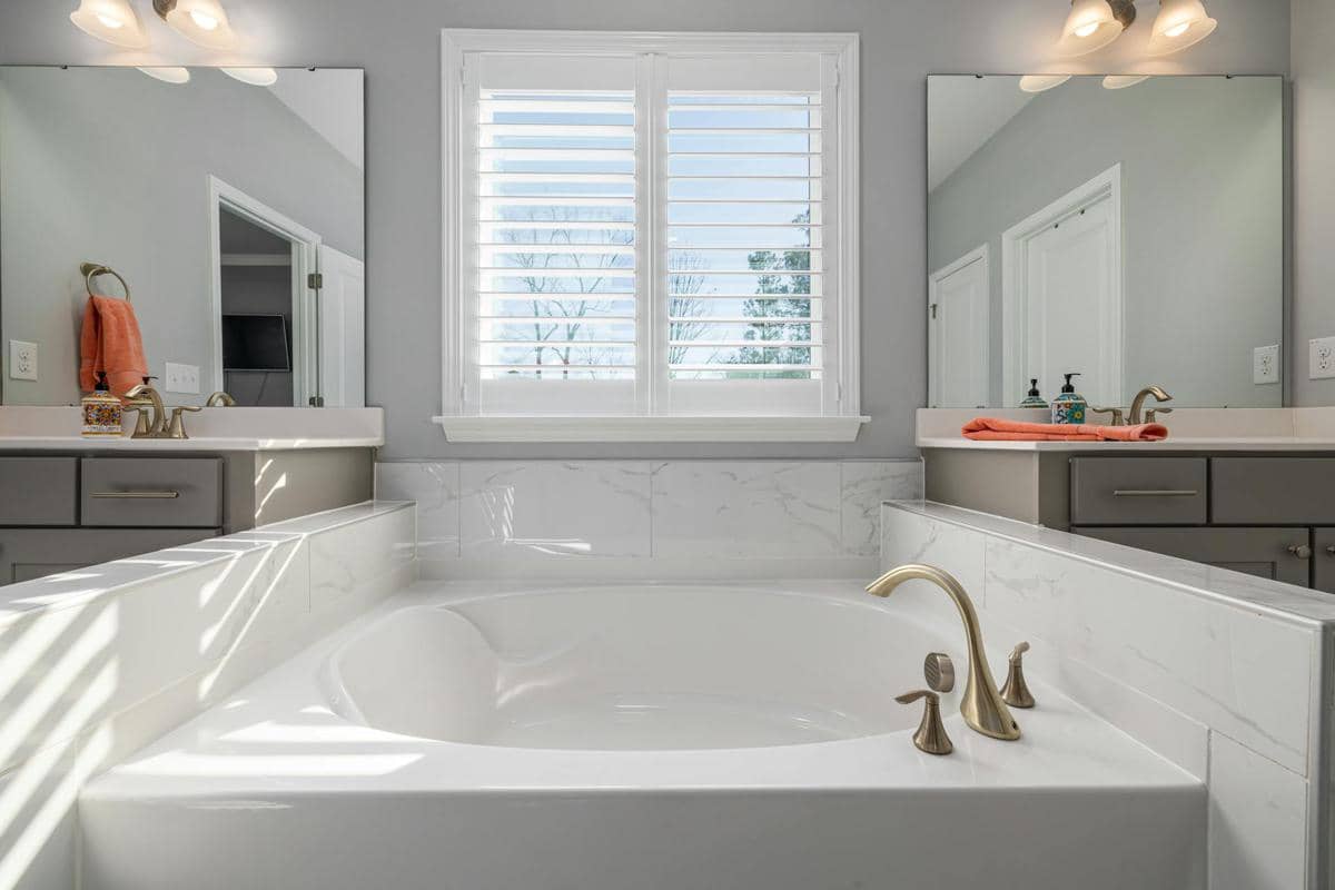 Kamar mandi modern dengan bathtub putih, jendela berjaluzi putih, dan wastafel ganda dengan perlengkapan berwarna emas.