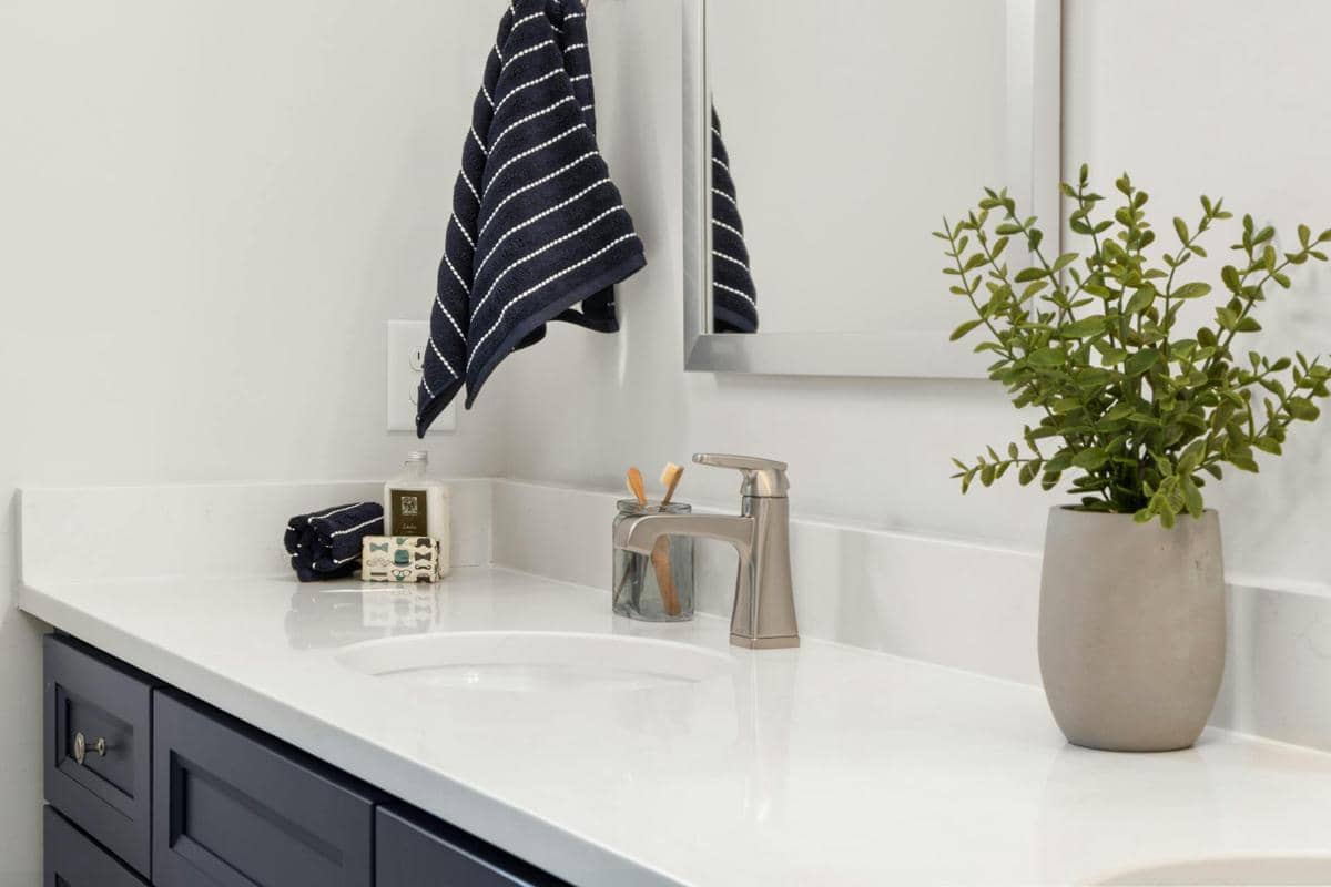 Kamar mandi modern dengan wastafel putih, keran logam, tanaman hias dalam pot abu-abu, dan handuk bergaris tergantung di dinding.