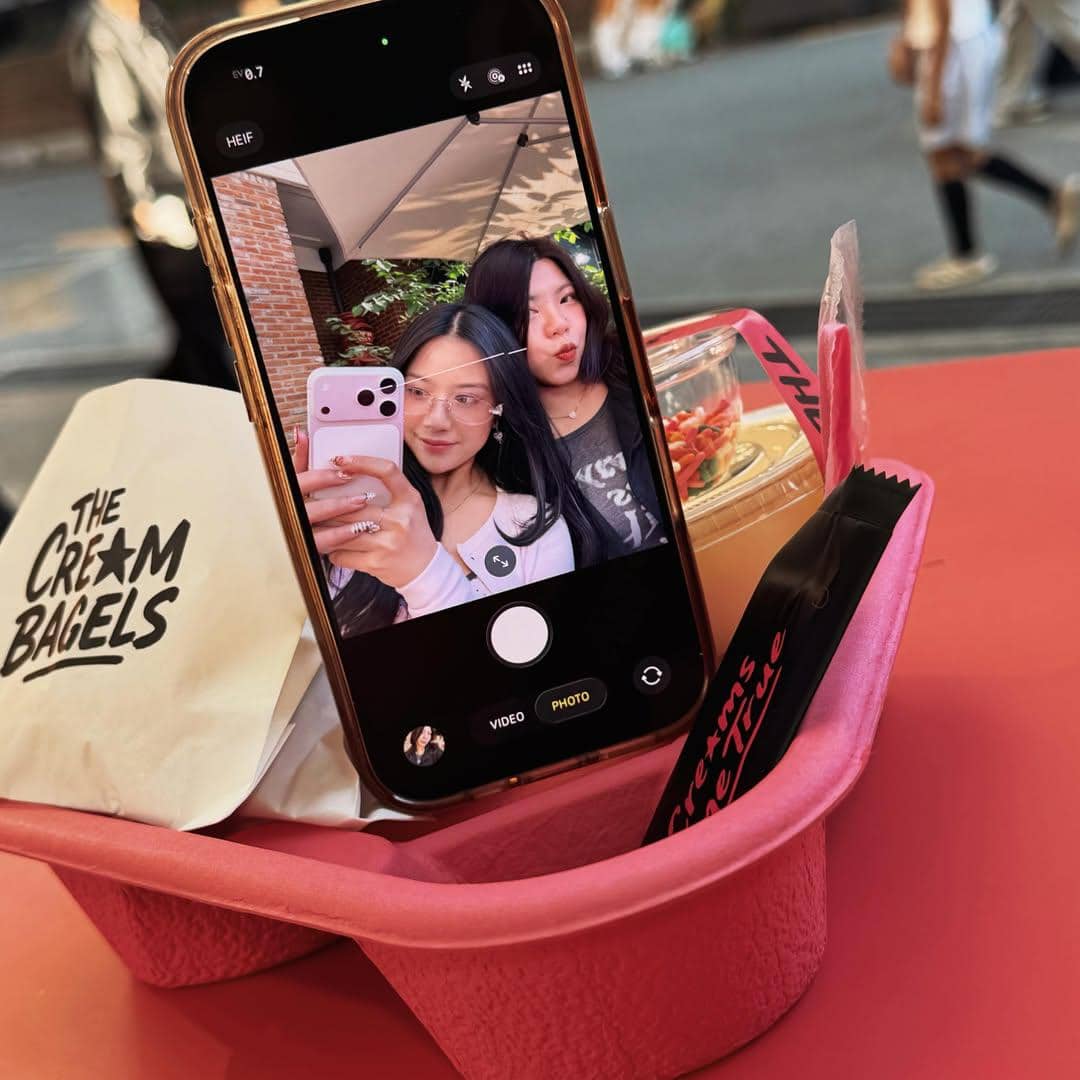 Dua perempuan berfoto selfie dengan ponsel di atas meja merah, di samping makanan penutup dan kemasan bertuliskan The Cream Bagels.