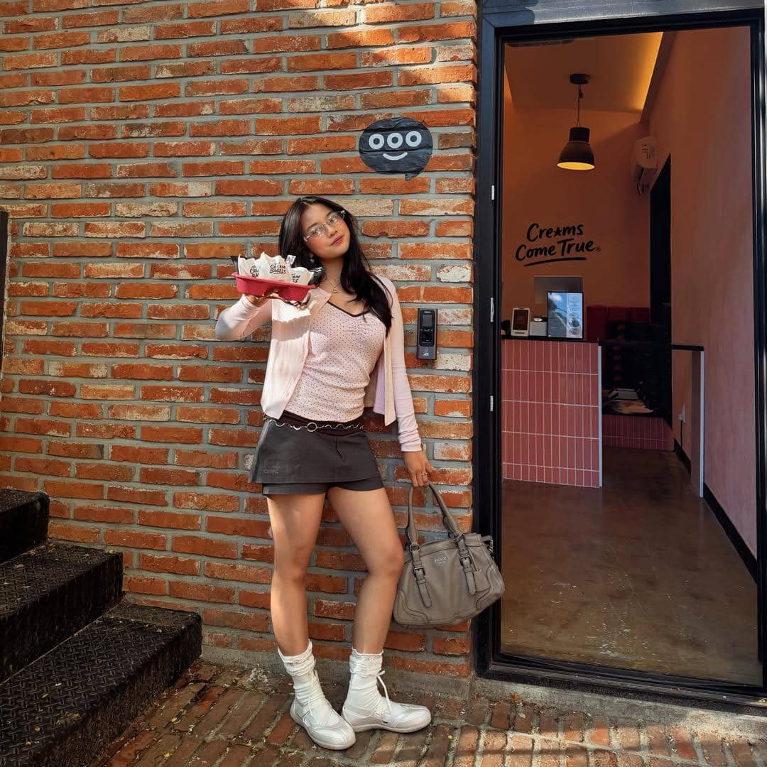 Seorang perempuan berpose di depan toko dessert di Seoul sambil memegang nampan berisi es krim dan tas tangan abu-abu.