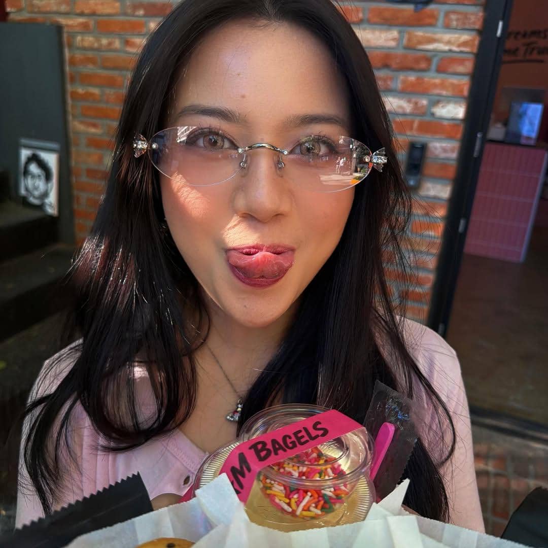 Seorang wanita berambut panjang mengenakan kacamata bening berpose menjulurkan lidah sambil memegang toples dessert bertuliskan bagels di Seoul.