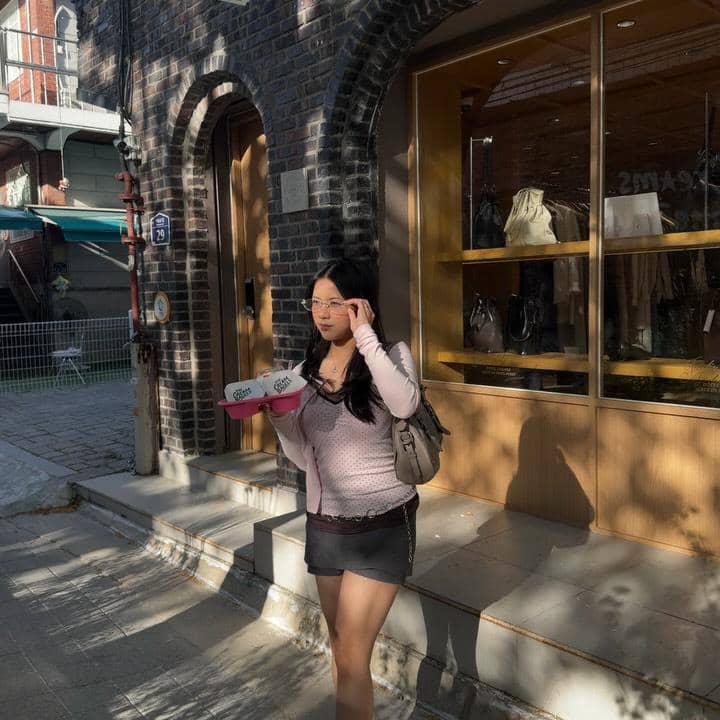 Seorang wanita mengenakan kacamata dan pakaian kasual membawa kotak dessert sambil berjalan di trotoar area pertokoan di Seoul.