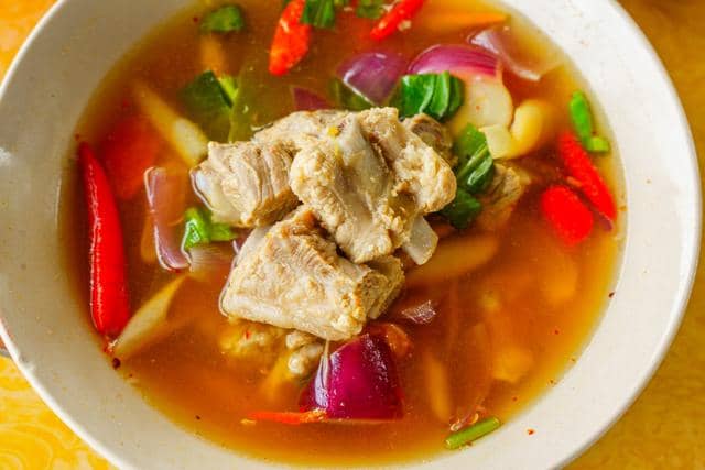 Semangkuk asem-asem daging dengan potongan daging sapi, cabai merah, bawang merah, dan sayuran dalam kuah bening berwarna kecokelatan.