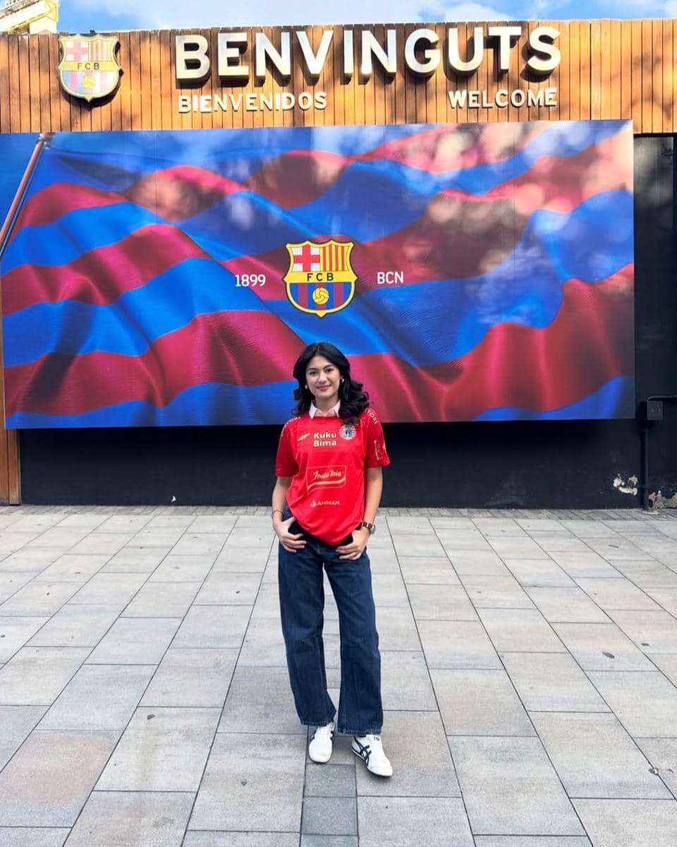 Seorang perempuan mengenakan kaus merah dan celana jeans berdiri di depan papan bertuliskan 'Benvinguts' dengan latar bendera klub sepak bola Barcelona.