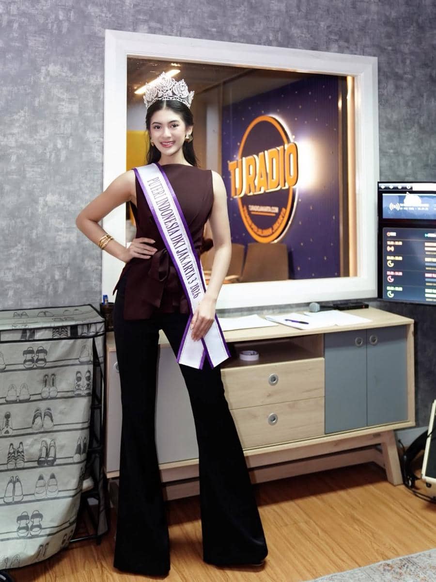 Seorang wanita mengenakan mahkota dan selempang kontes kecantikan berdiri di depan ruang siaran radio dengan latar bertuliskan U Radio.
