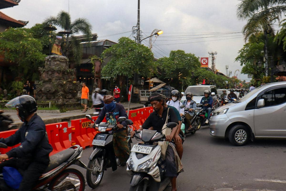 Pengendara motor dan mobil melintas di jalan kota yang padat dengan pembatas jalan oranye serta pepohonan di tepi jalan.