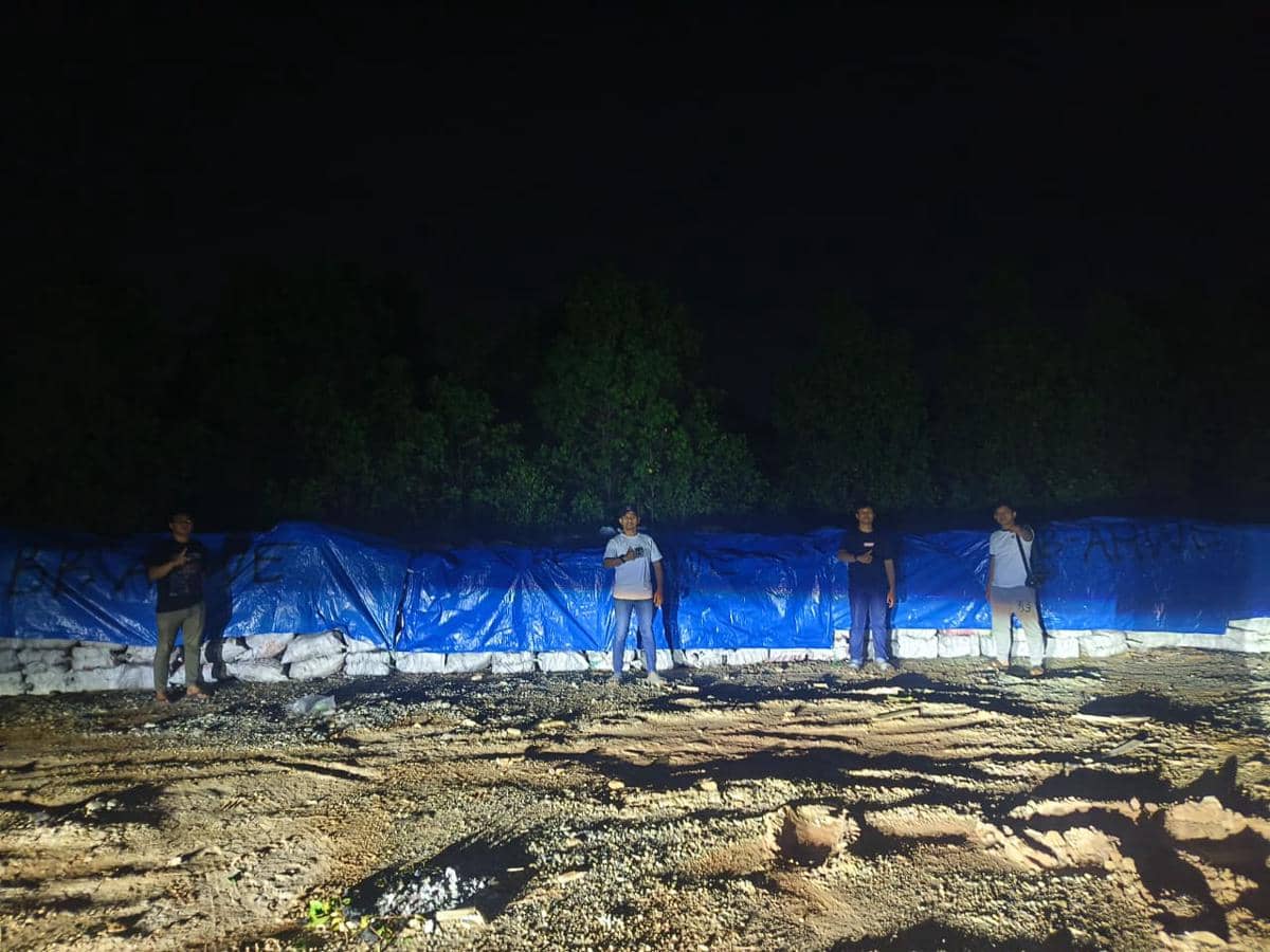 Beberapa petugas berdiri di depan tumpukan material yang ditutupi terpal biru pada malam hari di area hutan mangrove Kepulauan Meranti.