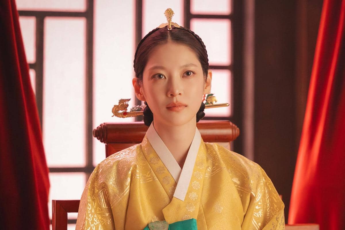 Seorang wanita mengenakan hanbok kerajaan berwarna emas dengan hiasan rambut tradisional duduk di kursi kayu berlatar tirai merah.