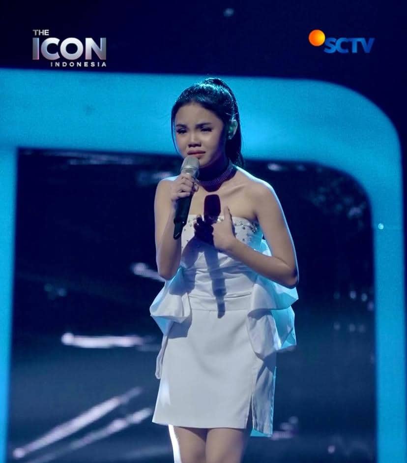Seorang peserta The Icon Indonesia mengenakan gaun putih sedang bernyanyi di atas panggung dengan pencahayaan biru dan mikrofon di tangan.