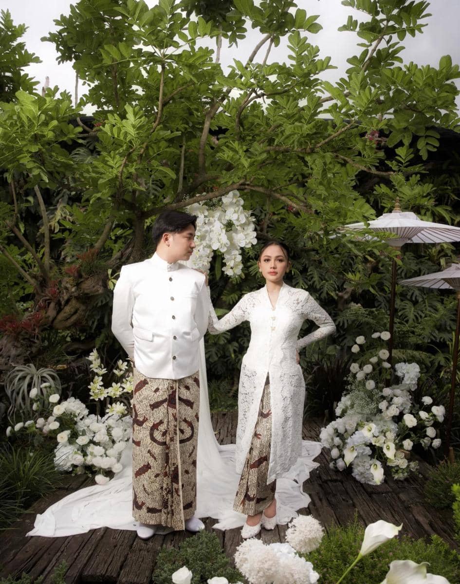Sepasang calon pengantin mengenakan busana adat Jawa berwarna putih dengan kain batik, berpose di taman penuh bunga dan pepohonan hijau.