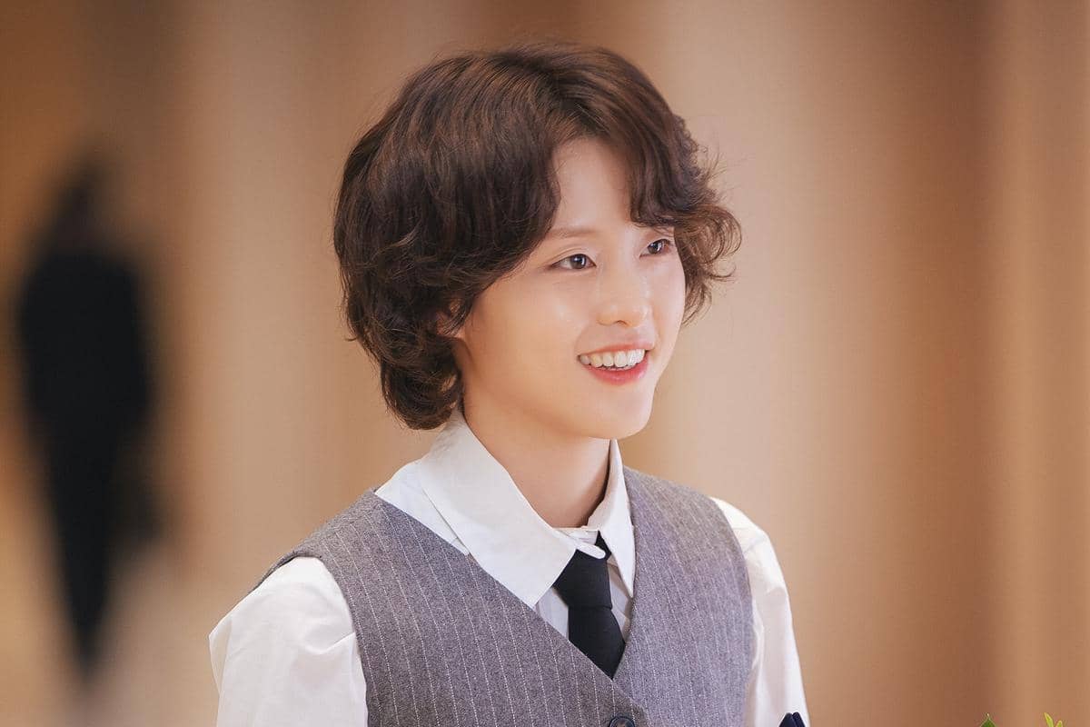 Seorang karakter tersenyum mengenakan kemeja putih, rompi abu-abu, dan dasi hitam dalam adegan drama Korea Perfect Crown.