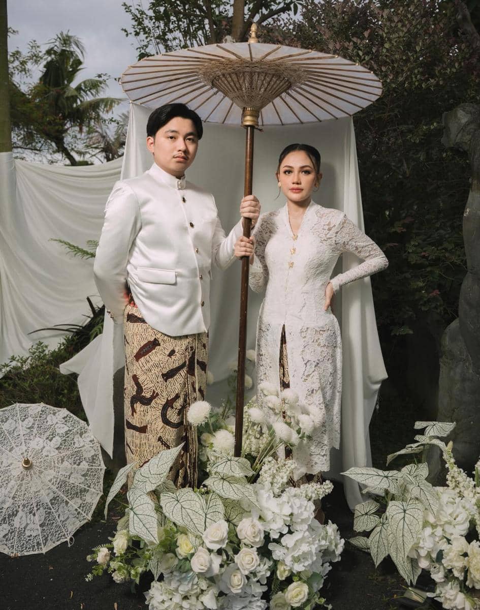 Sepasang calon pengantin mengenakan busana adat Jawa berwarna putih dengan latar dekorasi bunga dan payung tradisional di sesi prewedding.