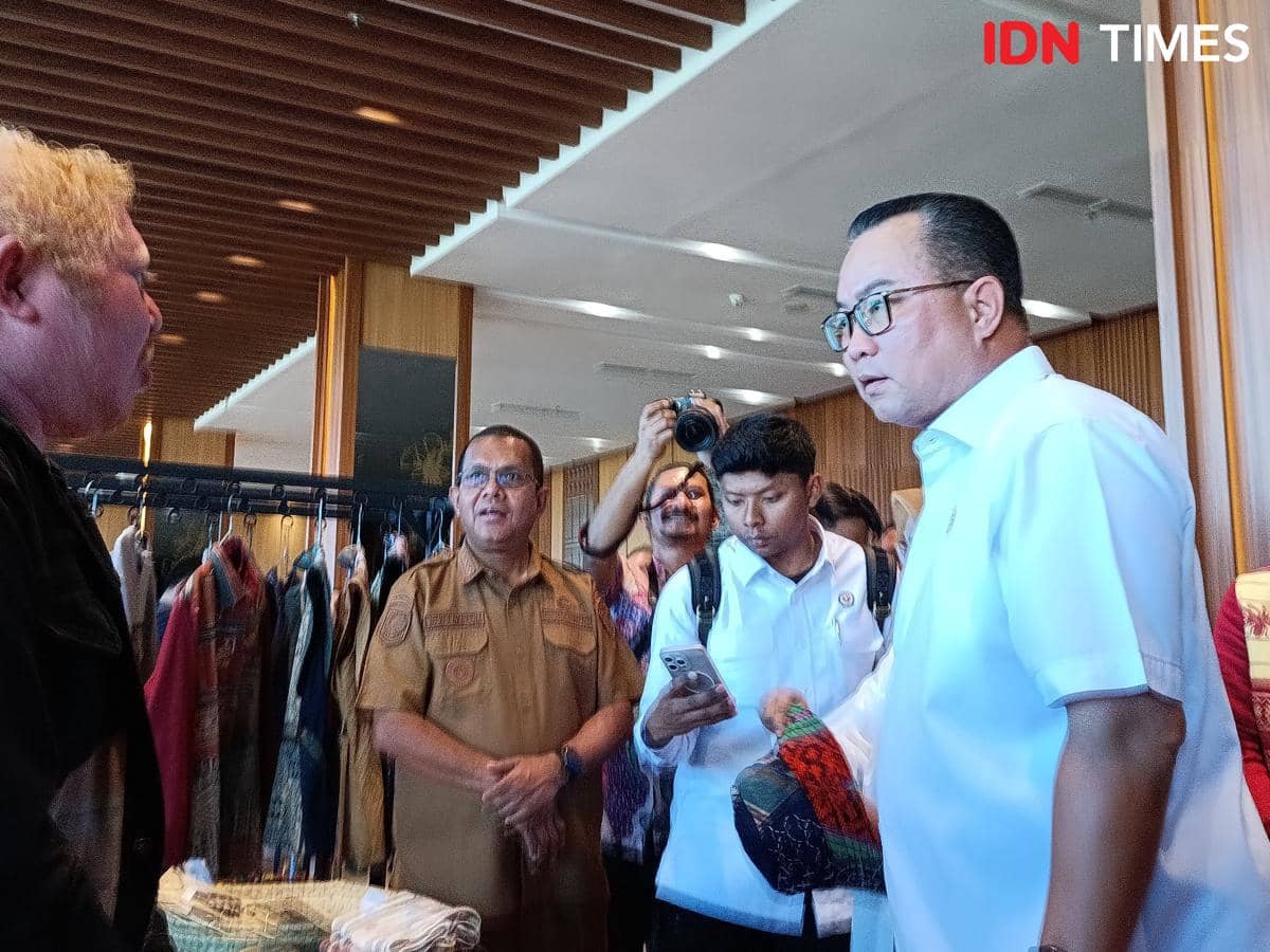 Seorang pria berbaju putih berbincang dengan beberapa orang di dalam ruangan dengan latar gantungan kain tradisional di Kupang, NTT.