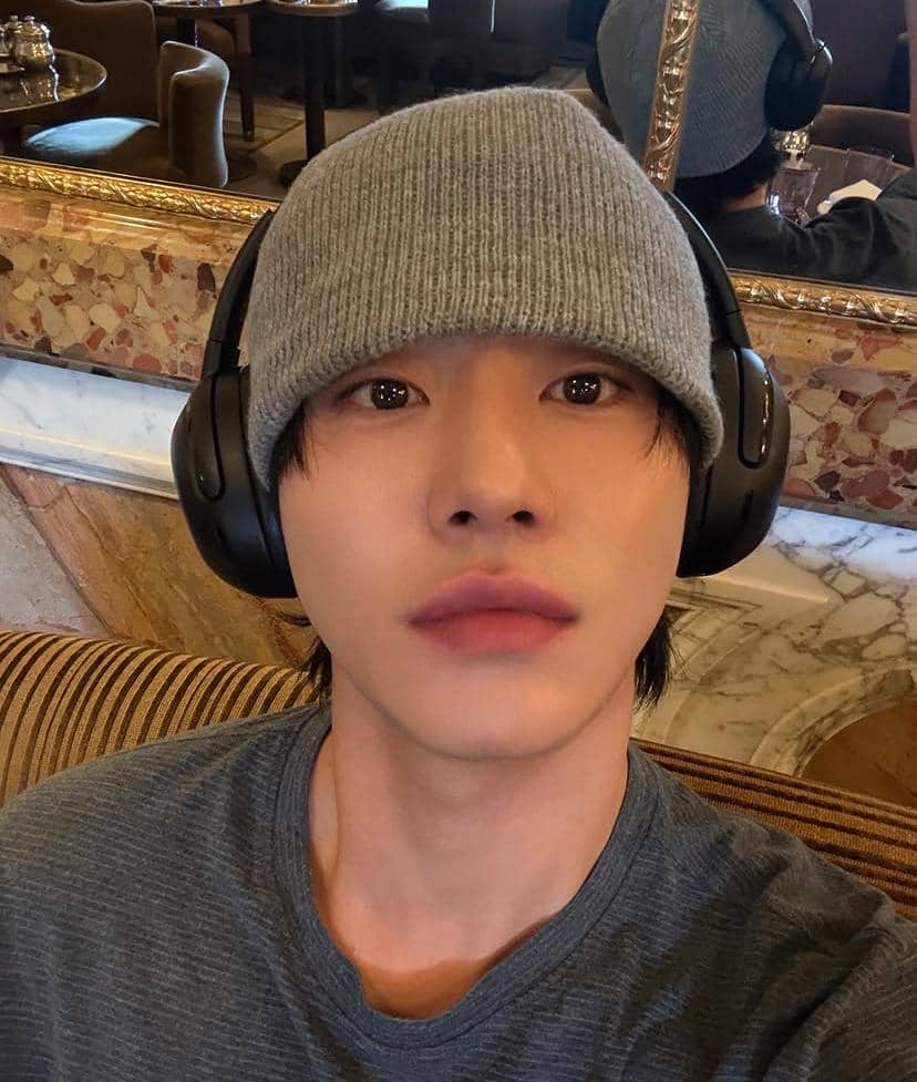 Seorang pria mengenakan beanie abu-abu dan headphone hitam berpose selfie di dalam ruangan dengan cermin besar di belakangnya.