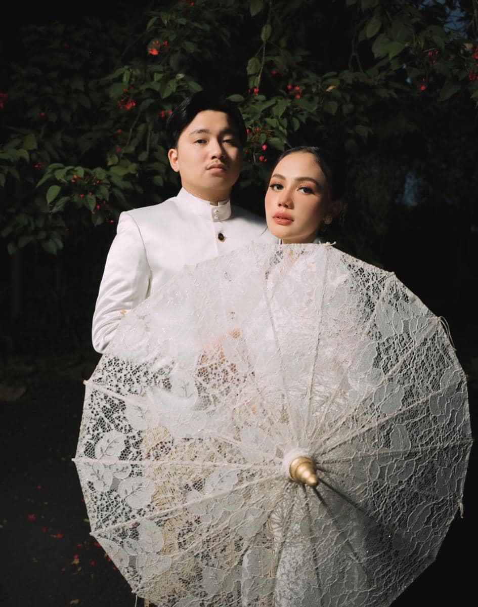 Sepasang calon pengantin berpose elegan dengan busana putih dan payung renda besar di depan latar pepohonan hijau.