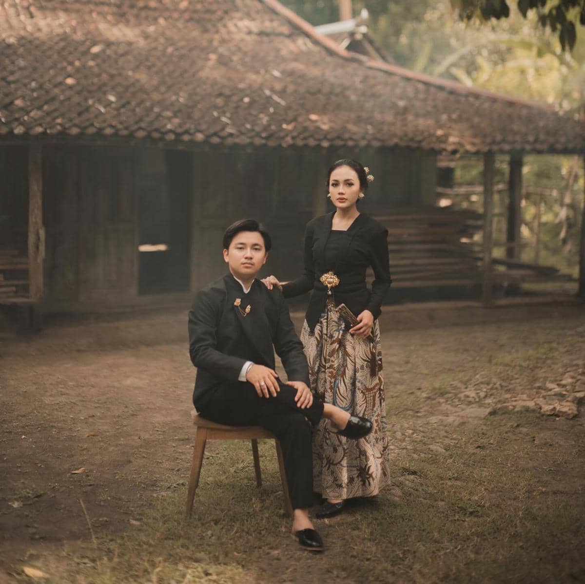 Sepasang calon pengantin berbusana adat Jawa berpose di depan rumah tradisional dengan suasana hangat dan klasik.