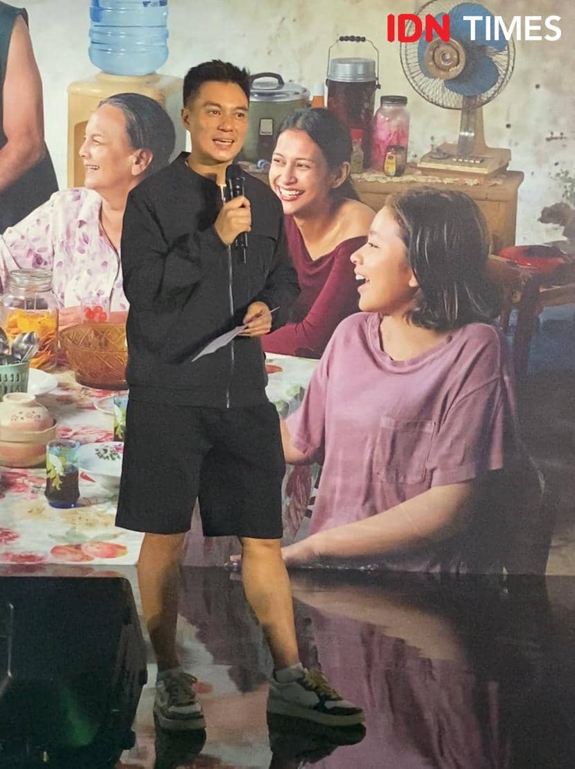 Seorang pria mengenakan pakaian hitam memegang mikrofon di acara gala premier film Semua Akan Baik-Baik Saja, dengan latar poster film.