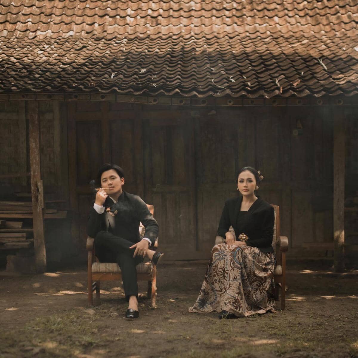 Sepasang calon pengantin duduk di kursi kayu mengenakan busana tradisional Jawa di depan rumah tua beratap genteng.