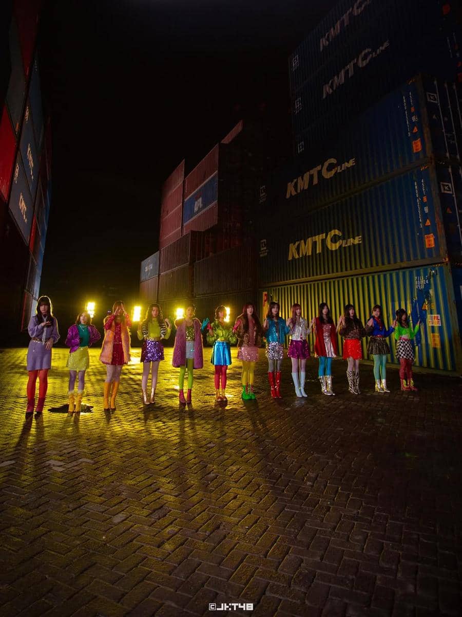 Belasan anggota grup idola perempuan berdiri berjajar mengenakan kostum warna-warni di area peti kemas pada malam hari.