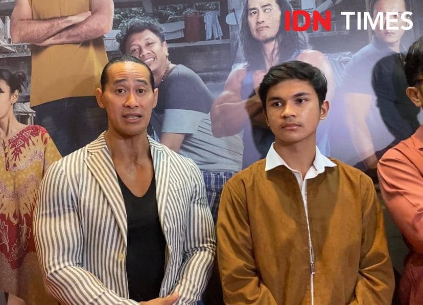 Dua pria berdiri berdampingan di acara press screening film Semua Akan Baik-Baik Saja, dengan latar poster film di belakang mereka.