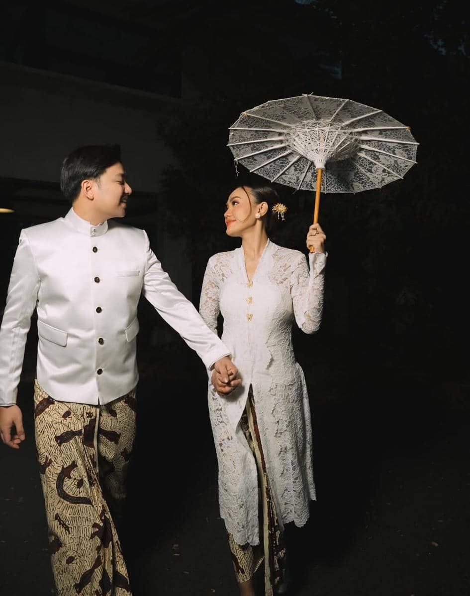 Sepasang calon pengantin mengenakan busana adat Jawa berwarna putih dan batik sambil bergandengan tangan di sesi foto prewedding.