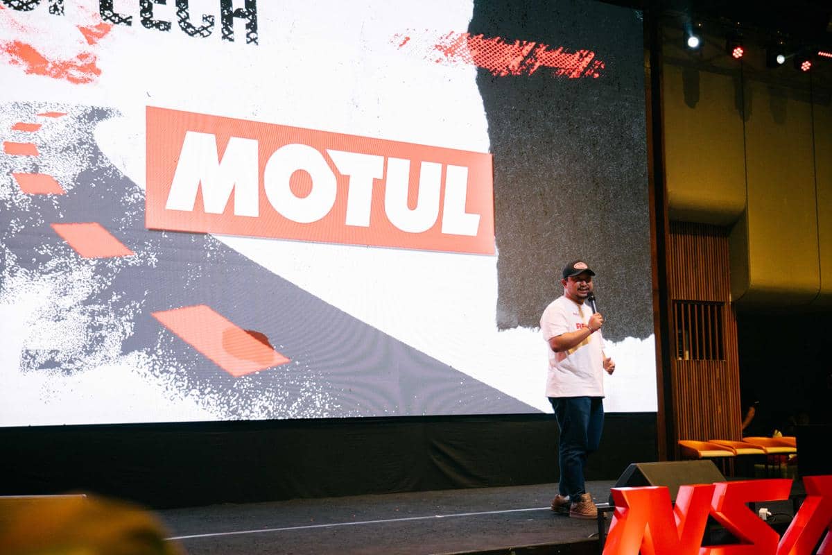 Seorang pembicara berdiri di atas panggung dengan latar belakang layar besar bertuliskan Motul dalam acara komunitas motor.
