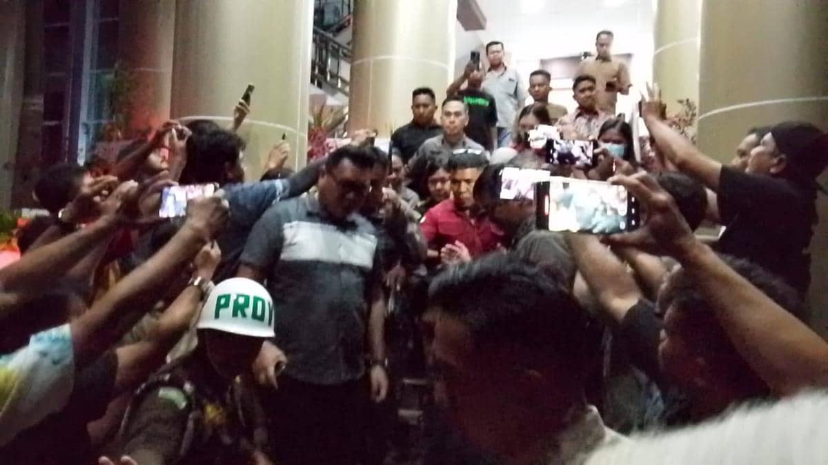 Kerumunan orang dan petugas mengawal seorang perempuan menuju mobil tahanan di halaman kantor Kejati Sulut pada malam hari.