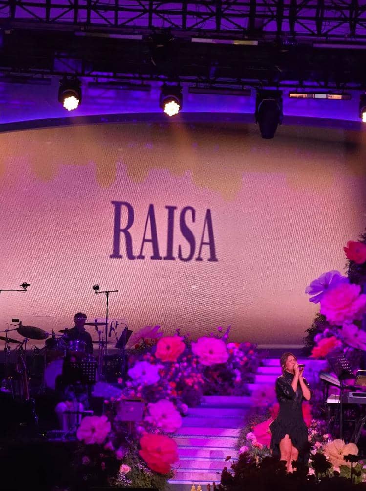 Penyanyi tampil di panggung konser dengan dekorasi bunga berwarna ungu dan merah muda, latar bertuliskan nama Raisa di layar besar.