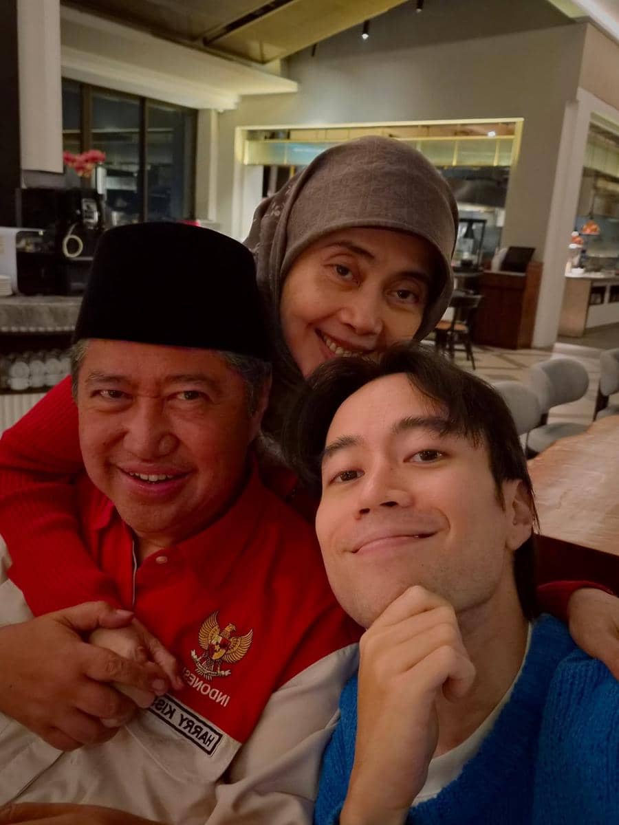 Seorang pria muda berpose bersama dua orang yang lebih tua, tersenyum hangat di dalam ruangan dengan pencahayaan lembut.