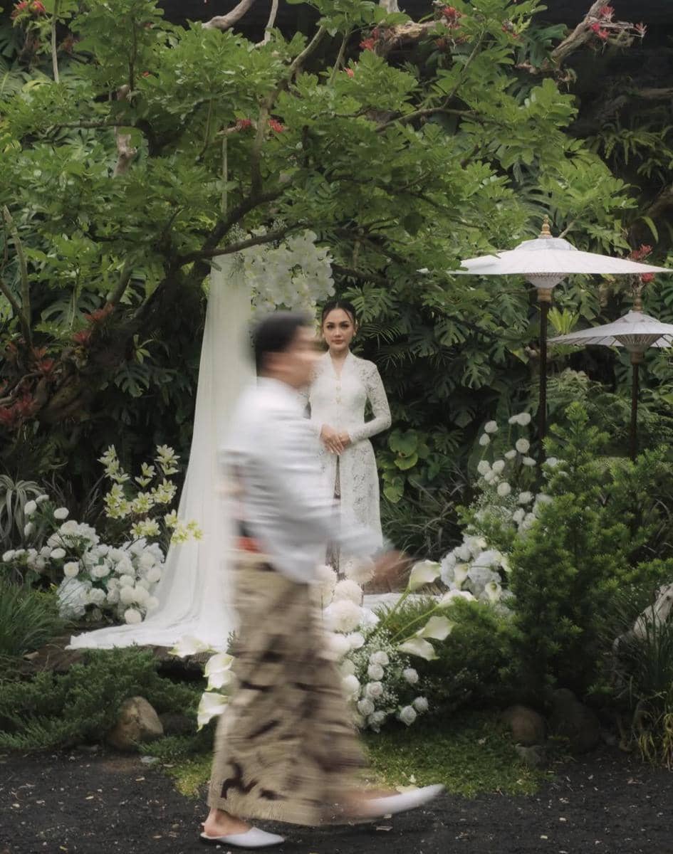 Seorang perempuan mengenakan kebaya putih berdiri di taman dengan dekorasi bunga putih, sementara seorang pria berjalan di depannya.