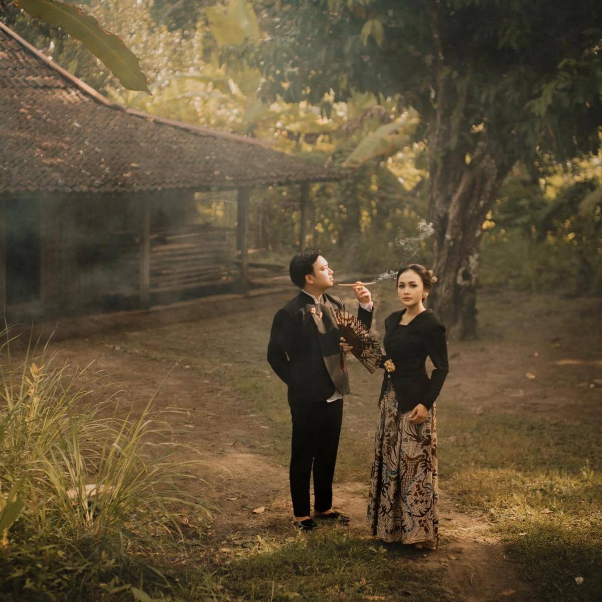 Pasangan mengenakan busana tradisional Jawa berpose di halaman rumah joglo dengan latar pepohonan dan suasana hangat sore hari.