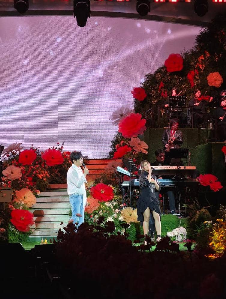 Dua penyanyi tampil di atas panggung konser dengan dekorasi bunga berwarna merah dan oranye, disertai pemain musik di latar belakang.
