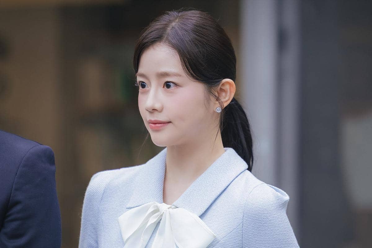 Seorang wanita berambut hitam dikuncir mengenakan blazer biru muda dengan pita putih dalam adegan drama Korea Perfect Crown.