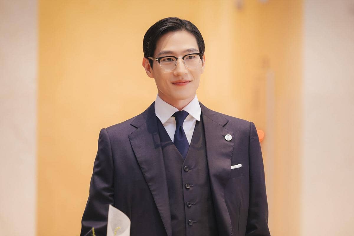 Seorang pria mengenakan setelan jas formal berwarna biru tua dan dasi, berdiri di ruangan dengan latar berwarna hangat.