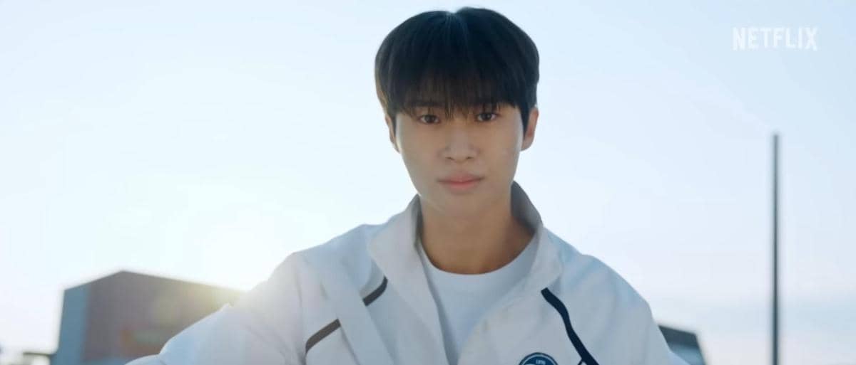 Seorang pria mengenakan jaket olahraga putih berdiri di luar ruangan dengan latar langit cerah dalam cuplikan serial Lovely Runner.